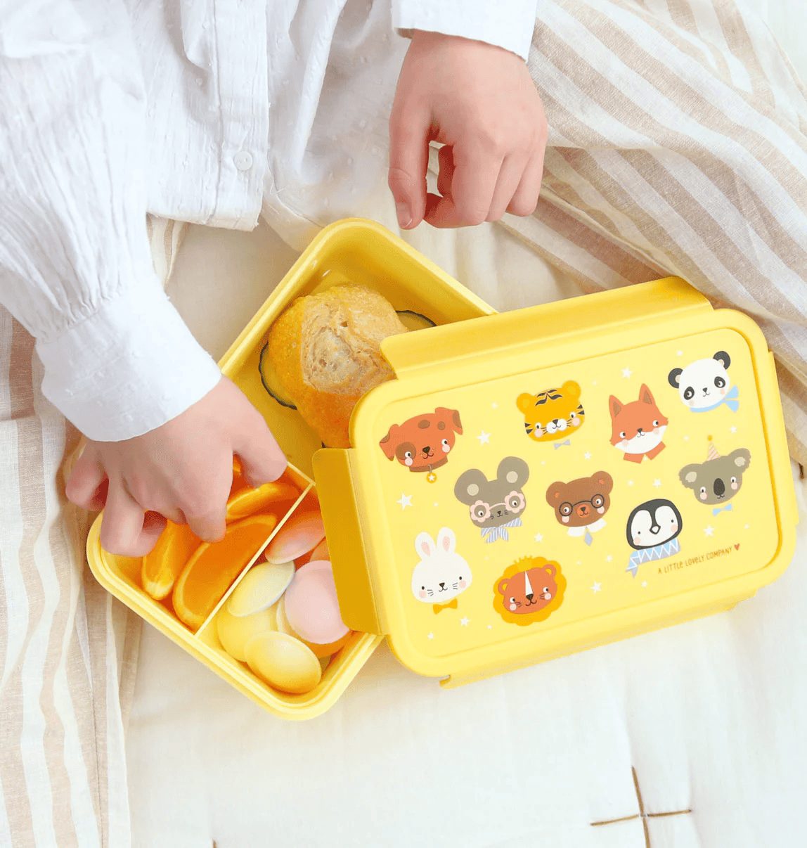A child’s hands are visible opening the lunch box, placing food inside the tray—demonstrating how the removable compartment works. The cheerful animal faces on the lid are visible in the background.