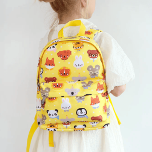 A child in a white dress wearing the cheerful bright yellow, patterned backpack by its top handle. The vibrant design includes playful animal faces including foxes, rabbits, bears and birds.