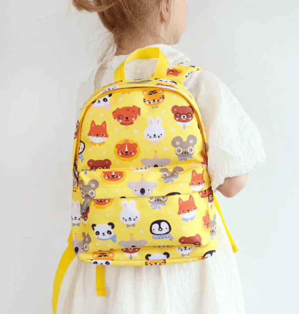 A child in a white dress wearing the cheerful bright yellow, patterned backpack by its top handle. The vibrant design includes playful animal faces including foxes, rabbits, bears and birds.