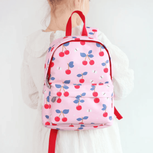 A child seen from behind wearing the pink cherry-patterned backpack. The side and front of the bag are visible, and the red-trimmed design contrasts with the child’s white outfit.