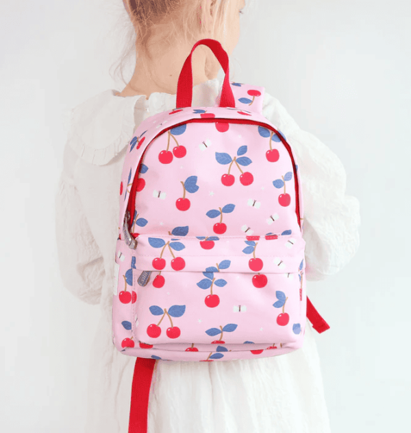 A child seen from behind wearing the pink cherry-patterned backpack. The side and front of the bag are visible, and the red-trimmed design contrasts with the child’s white outfit.