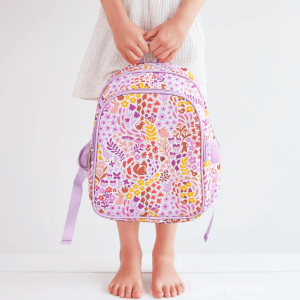 A child in a white dress stands barefoot on a white floor, holding the floral and woodland animal-themed backpack by its top handle. The vibrant design includes birds, squirrels, and flowers in shades of pink, yellow, and purple.