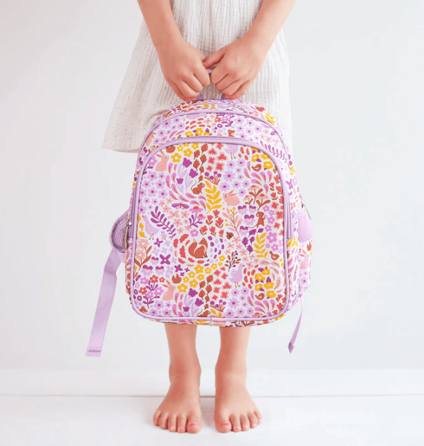 A child in a white dress stands barefoot on a white floor, holding the floral and woodland animal-themed backpack by its top handle. The vibrant design includes birds, squirrels, and flowers in shades of pink, yellow, and purple.