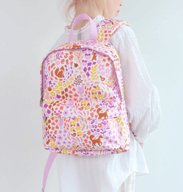 A child wearing the backpack, shown from the side against a neutral wall. You can see the bag’s main compartment and the floral‑fox print. The child’s white outfit highlights the pastel color palette of the design.