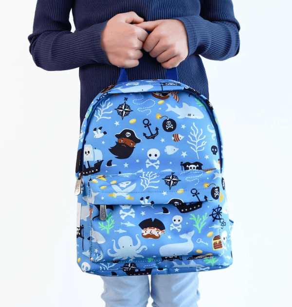 A child stands, holding the pirate‑design backpack by its top handle. The front pocket appear functional, and the playful print stands out against the kid’s outfit.